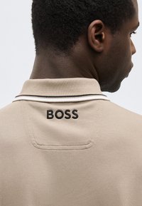 Άνδρας σε πλάγια θέση, φορώντας μπεζ πόλο πουκάμισο με κεντημένο το λογότυπο «BOSS» σε μαύρο χρώμα στη πάνω πλάτη κάτω από το γιακά.