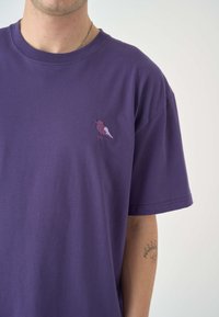 Lila Baumwoll-T-Shirt mit einem kleinen gestickten Vogelmotiv auf der linken Brust. Lockerer Schnitt mit kurzen Ärmeln und Rundhalsausschnitt.