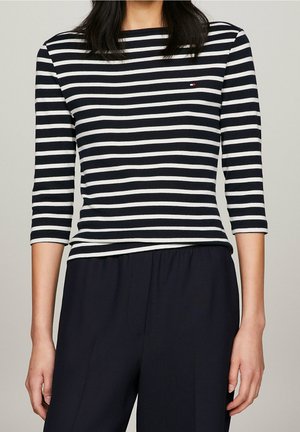 Femme portant un haut à manches longues rayé horizontal noir et blanc et un pantalon noir ample, debout devant un fond uni.