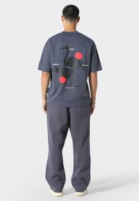 Grå bomulds t-shirt med korte ærmer med en sort kran-illustration og røde cirkler på ryggen. Løse grå bukser i matchende stil.