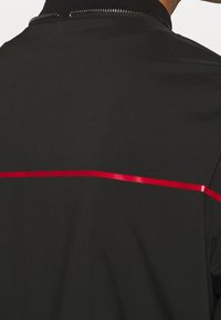 Zwarte jas met een gladde textuur, hoge kraag en een horizontale rode streep over de rug voor contrast.