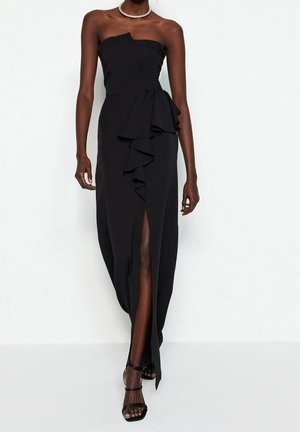 Femme portant une robe noire bustier avec un volant en cascade et une fente avant, assortie à des talons noirs à lanières et un collier de perles.