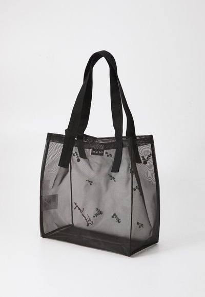 Transparent sort mesh tote bag med solide sorte stofhåndtag; har broderede grønne og røde blomster mønstre. Højden og bredden er synlige.