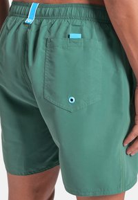 Costumi da bagno verdi con cintura elastica, tasca posteriore con etichetta blu e foro di drenaggio, indossati da una persona con braccia e mani visibili.