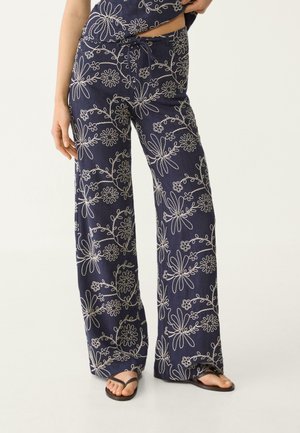 Marineblauwe broek met brede pijpen en witte bloemige borduur en een taille met trekkoord, gedragen met zwarte slippers op een lichte achtergrond.