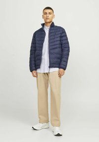 Marineblå vindjakke med horisontalt quiltet design, høj krave og lynlås, parret med en lys skjorte med knapper og beige bukser.