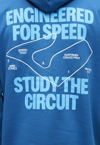 Μπλε ύφασμα με λευκή περίγραμμα του κυκλώματος του Αυστριακού Γκραν Πρι, με επισημασμένες ζώνες και κείμενο «Σχεδιασμένο για ταχύτητα» και «Μελετήστε το Κύκλωμα».