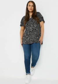 Blusa nera con motivo floreale, maniche corte a volant e dettaglio a chiave sullo scollo, abbinata a jeans blu e sneakers bianche.