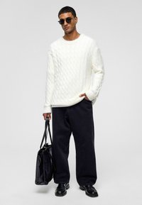 Flauschiger weißer Pullover mit Kabelmuster, kombiniert mit weiten schwarzen Hosen. Accessoires sind schwarze Sonnenbrillen und eine strukturierte schwarze Tasche.