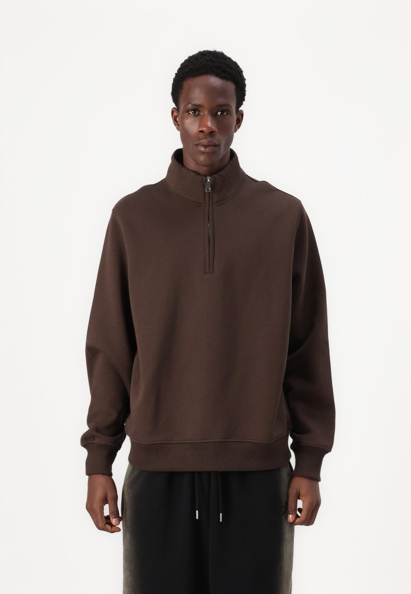 Brun, højhalset sweatshirt med en halv lynlås; afslappet pasform, blød tekstur, ribbede manchetter og kant, båret over mørke, løstsiddende bukser.