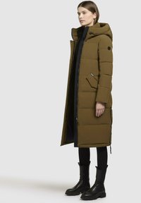 Olive green puffer coat with a hood, black zipper, and pockets. Features a quilted texture and slightly oversized fit, paired with black boots.