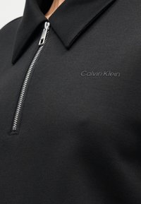 Μαύρο πόλο μπλούζα με φερμουάρ, κατασκευασμένη από λείο ύφασμα. Διαθέτει μυτερό γιακά και ασημί φερμουάρ. Η επιγραφή "Calvin Klein" είναι ανάγλυφη στο πουκάμισο.