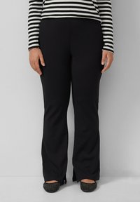 Schwarze, maßgeschneiderte Hose mit weitem Schnitt, aus glattem Stoff. Mit seitlichen Schlitzen am Saum. Kombiniert mit schwarzen, verzierten Ballerinas.