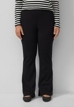 Zwarte, op maat gemaakte broek met een uitlopende stijl, gemaakt van een gladde stof. Heeft zijsplits aan de zoom. Gecombineerd met zwarte versierde flats.