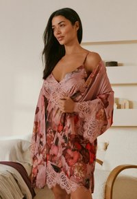 Femeie purtând o cămașă de noapte satinate roz cu dantelă florală și un halat asortat, stând într-un dormitor minimalist, iluminat delicat.