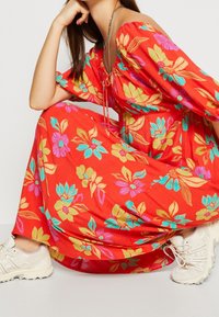 Rotes Maxikleid mit floralen Mustern in Gelb, Pink und Aquamarin. Verfügt über schulterfreie Ärmel und einen Kordelzug in der Taille. Kombiniert mit weißen Sneakern.