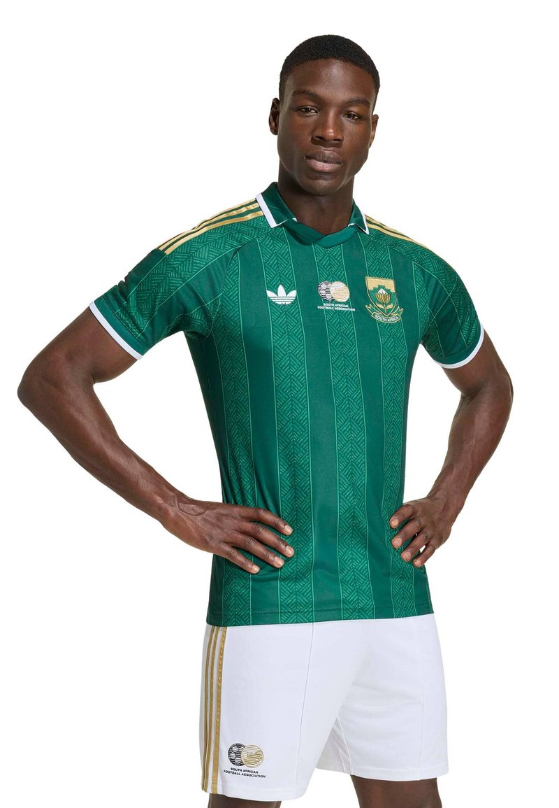 Homme portant un maillot de football sud-africain vert et un short blanc, les mains sur les hanches, debout devant un fond blanc.