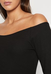 Haut noir côtelé à épaules dénudées et manches longues, avec un décolleté large et un tissu texturé pour plus de détails.