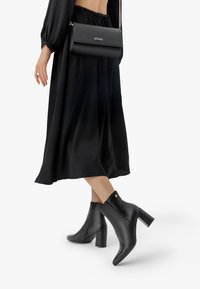 Mustat nilkkurit korkean tukikorkean, sileä nahkapinta, takana vetoketju ja koristeellinen kultasävy. Yhdistettynä leijaavaan mustaan mekkoon.