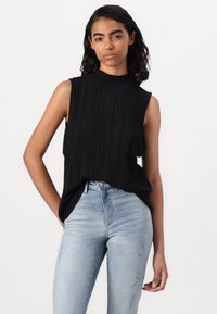 Haut sans manches noir à col montant, orné de plis verticaux, associé à un jean skinny délavé avec des accents déchirés.