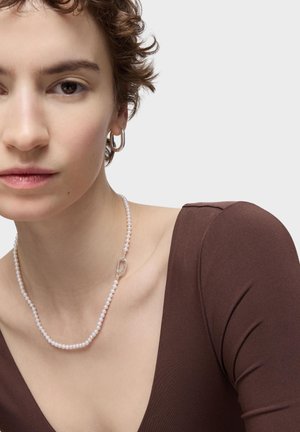 Primo piano di una persona con capelli corti e ricci che indossa una maglia marrone, una collana di perle con chiusura in argento e un orecchino a cerchio in argento.