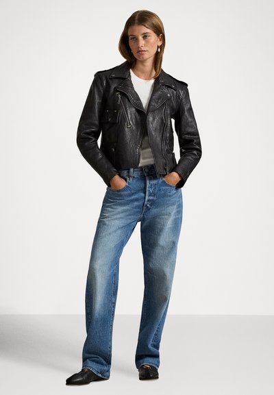 Crna kožna motoristička jakna s džepovima na patentni zatvarač, uparena s izblijedjelim plavim trapericama širokih nogavica. Model stoji s rukama u džepovima, noseći crne cipele.