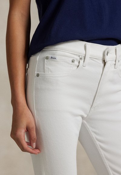 Osoba ubrana w białe dżinsy z denimu i granatową bluzkę, pokazująca przednią kieszeń dżinsów oraz etykietę marki Polo.