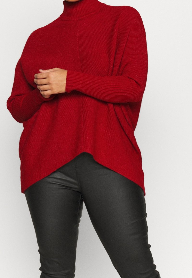 Femme portant un pull col roulé rouge ample à manches longues et un pantalon en cuir noir ajusté, debout devant un fond uni.