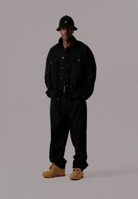 Čierna denimová bunda a nohavice. Biela košeľa pod ňou. Čierny vedierkový klobúk a hnedé pracovné topánky. Neformálny strih s gombíkovými detailmi.