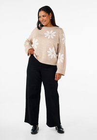 Beige gestrickter Pullover mit weißem Blumenmuster, weitem Ärmeln und rundem Ausschnitt, kombiniert mit schwarzen weiten Hosen und schwarzen Schuhen.