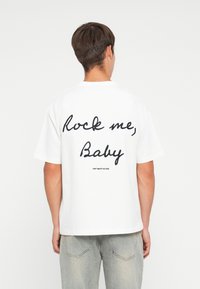Witte katoenen t-shirt met korte mouwen, voorzien van grote zwarte cursieve tekst op de achterkant met de tekst "Rock me, Baby" en een klein logo eronder.