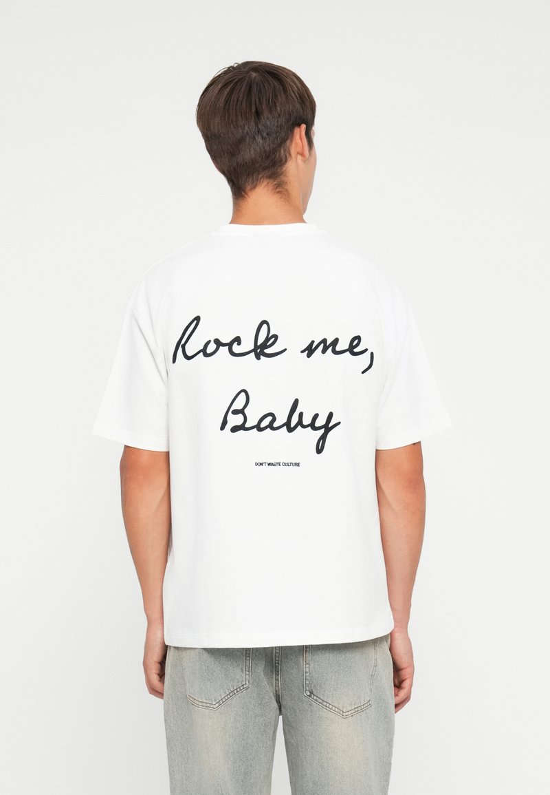 T-shirt en coton blanc à manches courtes, avec un grand texte noir en cursive au dos indiquant "Rock me, Baby" et un petit logo en dessous.