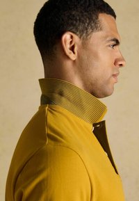 Kollane polosärk, mille krael on triibud. Krae sisaldab rohelist aktsenti. Kangas on sileda tekstuuriga ja sobib hästi peale.