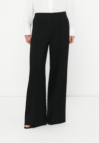 Woman wearing black wide-leg trousers and a white long-sleeve shirt, standing on a white floor against a white wall.