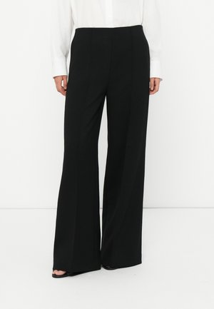 Woman wearing black wide-leg trousers and a white long-sleeve shirt, standing on a white floor against a white wall.