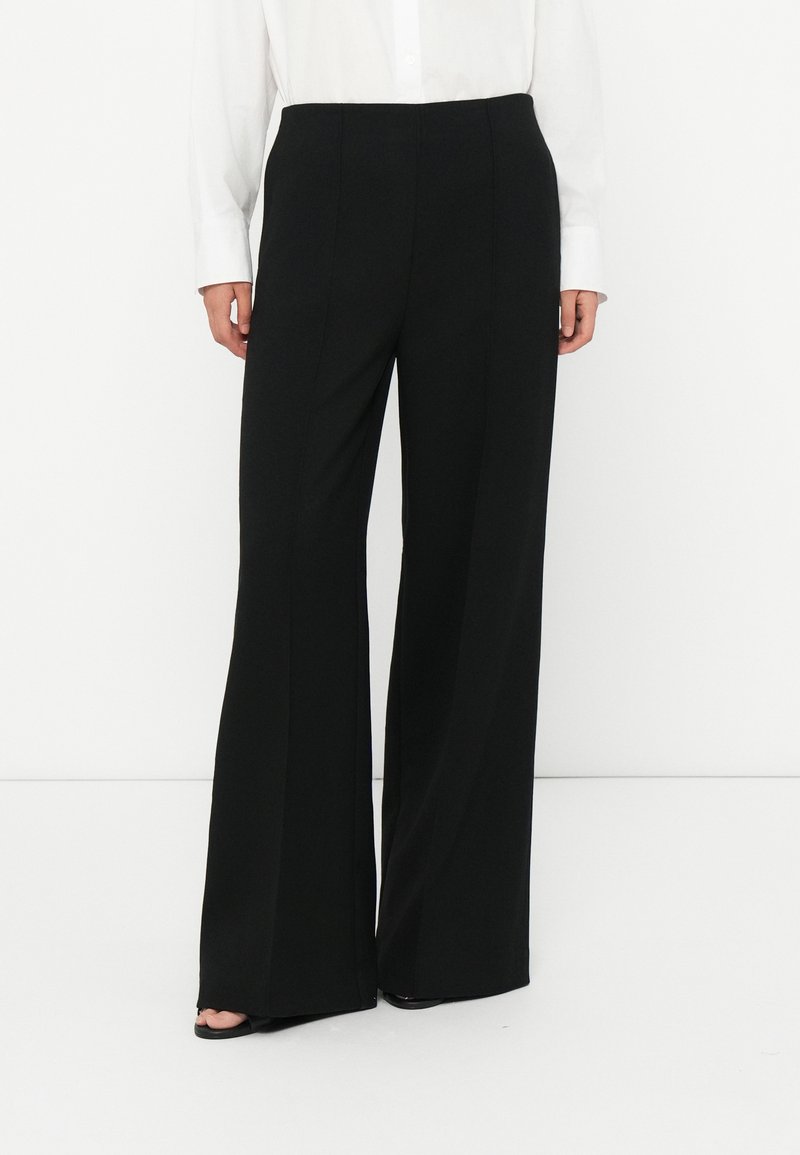 Woman wearing black wide-leg trousers and a white long-sleeve shirt, standing on a white floor against a white wall.
