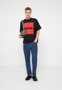 Crna majica s crvenim kvadratnim grafičkim dizajnom i tekstom 'Calvin', u kombinaciji s plavim trapericama ravnog kroja i crnim cipelama, nosi sivu torbu preko ramena.