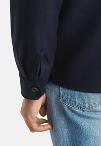 Camicia blu navy con tessuto testurizzato, dotata di dettagli con polsini abbottonati e una leggera apertura, abbinata a jeans in denim chiaro.