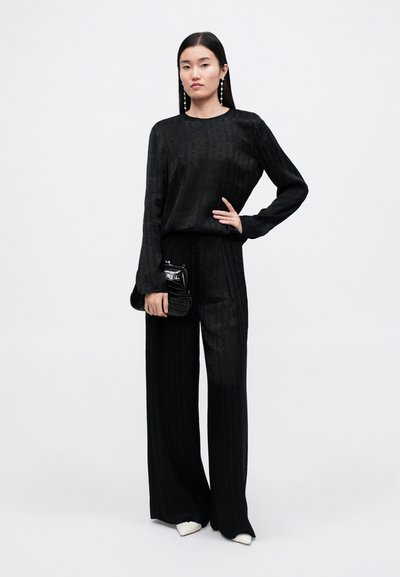Dvojdielny čierny outfit s dlhým rukávom a širokými nohavicami, s textúrovaným vzorom. Modelka drží malú čiernu listovku.