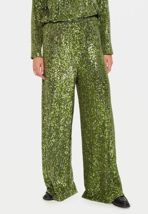 Woman wearing green sequined wide-leg pants paired with black shoes, standing against a plain light background.