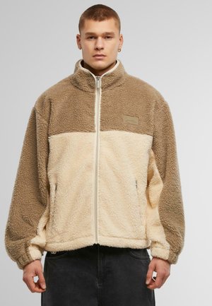 Fleece jacket in light beige and brown, featuring a high collar, zip closure, and two side pockets. Soft, textured fabric with a sporty design.