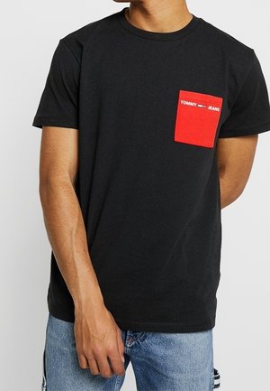 Mann in einem schwarzen T-Shirt mit kurzen Ärmeln und roter Brusttasche mit der Aufschrift "Tommy Jeans" sowie hellblauen Jeans, der vor einem weißen Hintergrund steht.