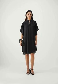 Μαύρο φόρεμα μέχρι το γόνατο με φουσκωτά μανίκια και λαιμόκοψη σε σχήμα κλειδώματος. Συνδυάζεται με μαύρη τσάντα χειρός και γόβες με λουράκι στον αστράγαλο. Λείο υφασμάτινο υλικό.