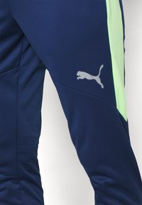 Marineblauwe sportbroek met een contrasterend lichtgroen paneel, gemaakt van gladde stof, met een reflecterende grijze Puma-logo op de dij.