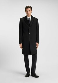 Cappotto lungo in lana nera con un design su misura, dotato di chiusura con bottone a un solo petto, due tasche laterali e rever a scollatura, abbinato a pantaloni scuri.