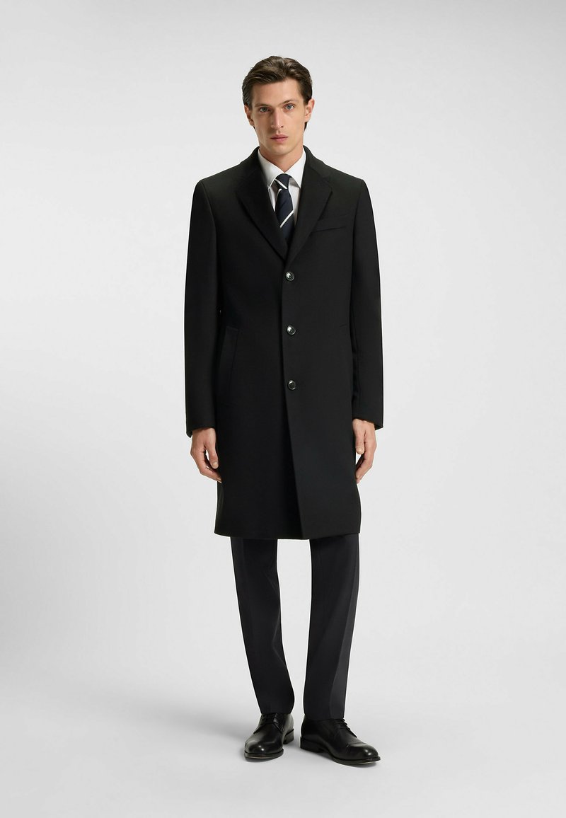 Cappotto lungo in lana nera con un design su misura, dotato di chiusura con bottone a un solo petto, due tasche laterali e rever a scollatura, abbinato a pantaloni scuri.