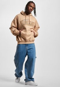 Bežinė gobtuvinė maikutė su priekinėmis kišenėmis ir logo, derinama su šviesiai mėlynais plačiais džinsais ir baltais sportbačiais. Sklandus audinys, kasdienis dizainas.