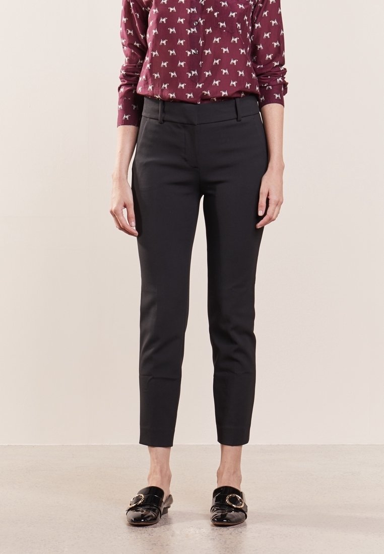 Femme portant un pantalon noir slim longueur cheville, un haut à manches longues bordeaux avec un motif de chien blanc, et des chaussures noires sans lacets avec des boucles dorées.
