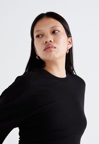Black long-sleeve top with a close fit and round neckline, worn with gold hoop earrings. Smooth texture, minimal design.