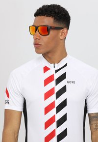 GORE sykkeltopp har et hvitt stoff med røde og svarte diagonale striper, korte ermer og glidelås foran. Sorte solbriller med oransje linser.